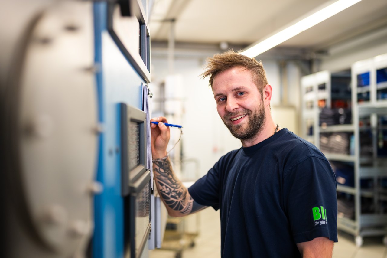 Ein lächelnder Mann mit Bart und tätowiertem Arm schreibt mit einem Stift an einer blauen Maschine.
