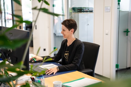 Lächelnde Frau mit kurzen Haaren sitzt am Schreibtisch vor einem Computer und arbeitet.