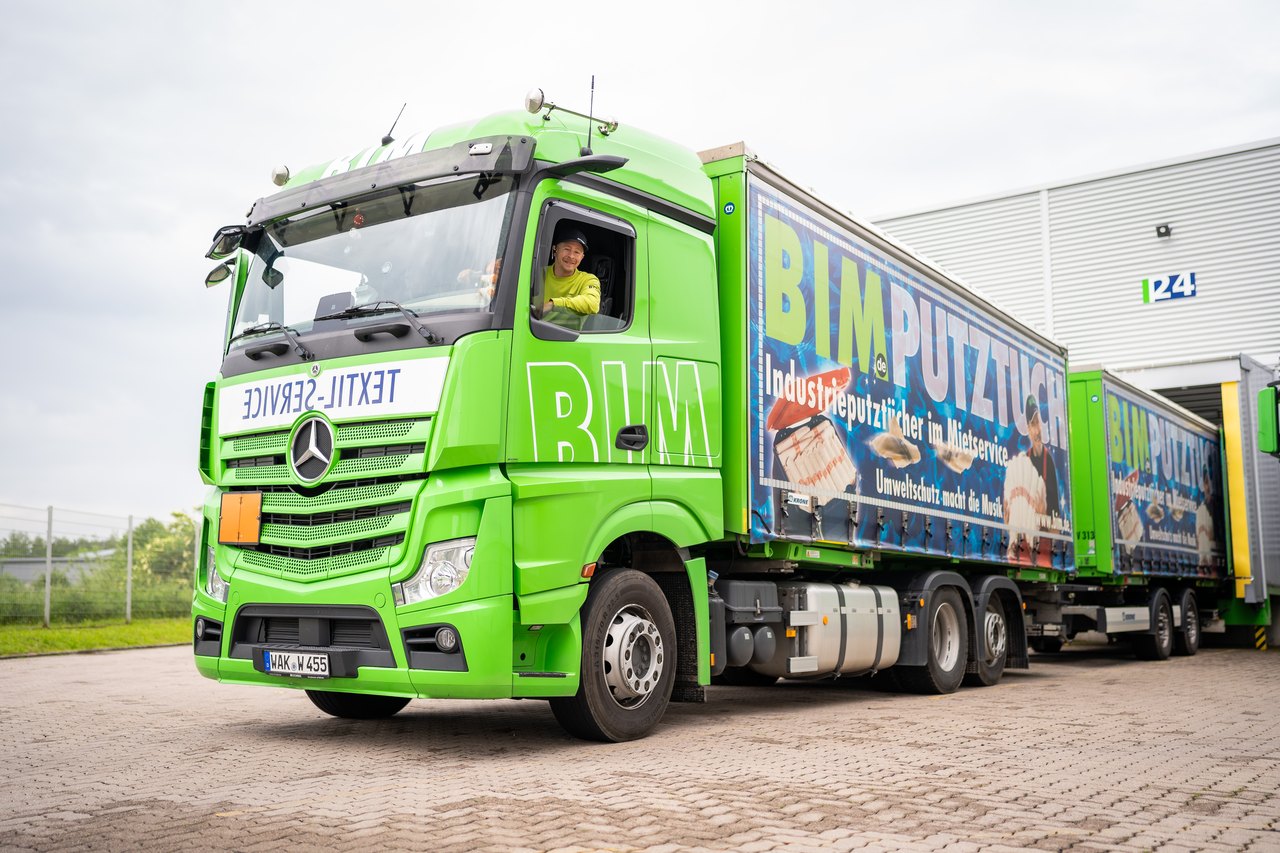 Grüner Mercedes-LKW mit Anhänger, beschriftet mit "BIM PUTZTUCH" und "TEXTIL SERVICE", Fahrer im Führerhaus.